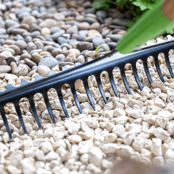 A sleek Double Sided Soil Rake - Carbon Steel rests on a bed of small stones and pebbles, its FSC Certified Ash Wood handle partially visible in the foreground.
