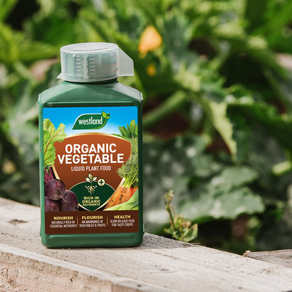 A bottle of Westland Organic Vegetable Liquid Plant Food 1L, designed to enhance nutrient availability for optimal vegetable growth, is placed on a wooden surface with lush plants in the background.
